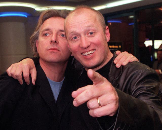 Rik Mayall, left, with his left hand on Adrian Edmondson's shoulder. Edmondson has his right hand on Mayall's shoulder and is smiling and pointing at the camera with his left hand