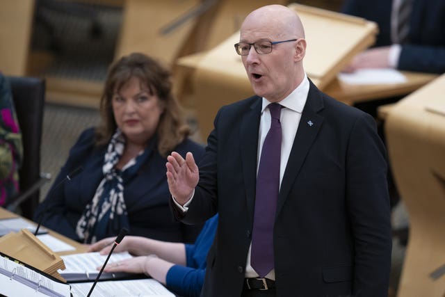 John Swinney standing while speaking in Parliament, with Fiona Hyslop seated beside him