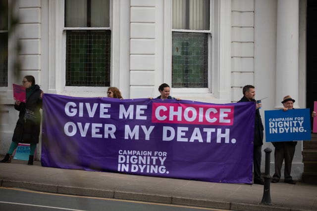 A group of people stand on a pavement holding a purple banner that reads 'give me choice over my death'