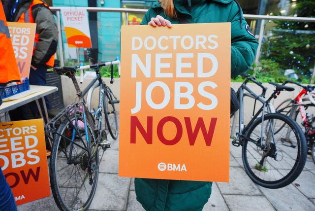A woman on a picket line holding a placard which reads 'Doctors need jobs now'