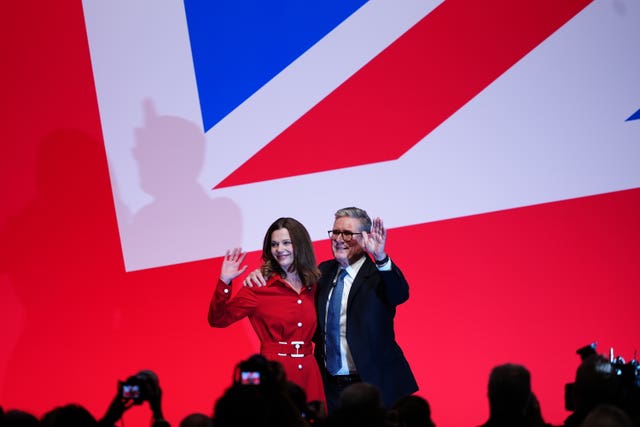 Sir Keir and Victoria Starmer wave on stage, with a large British flag in the background