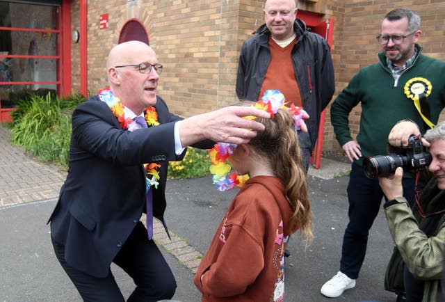 Scottish First Minister and SNP leader John Swinney (left) presents a lei (a garland or necklace of flowers given in Hawaii as a token of welcome or farewell) to one of the children, as SNP parliamentary candidate for Glasgow South Stewart McDonald (right) looks on, during a visit to the Jeely Piece Club in Glasgow