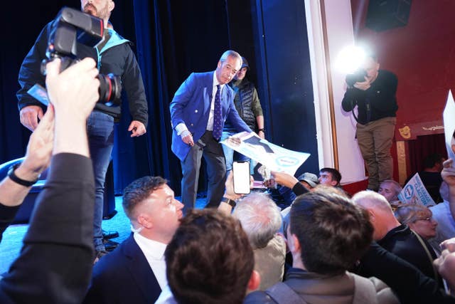 Nigel Farage stands on a stage accepting posters from the audience below as a photographer takes his picture 
