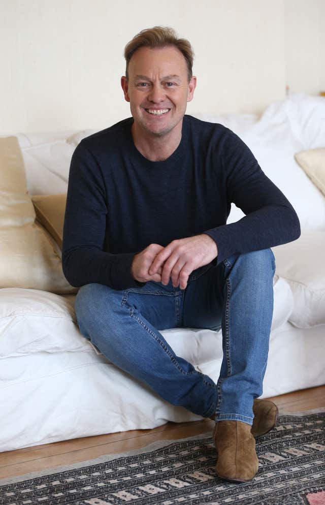 Jason Donovan sitting down and smiling while wearing a black top, blue jeans and brown shoes