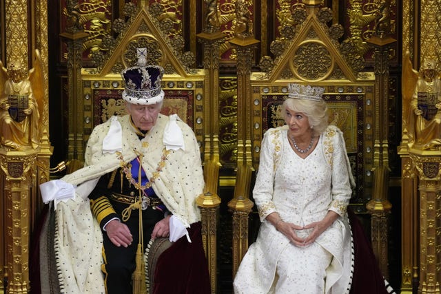 Charles and Camilla on thrones in the House of Lords with the King wearing ermine and his crown and Camilla in a white gown sitting beside him 