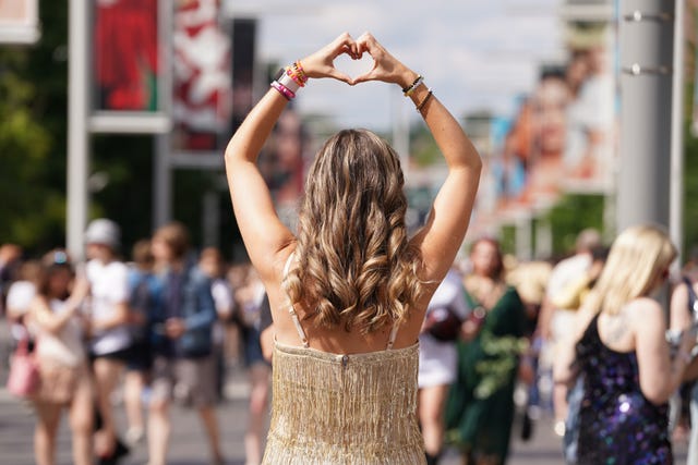 A Taylor Swift fan, back to camera, raises her arms and make a heart symbol with her hands