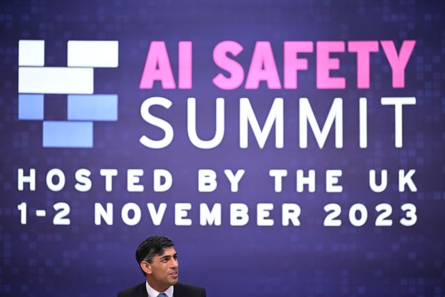 Rishi Sunak sits below a large sign reading 'AI Safety Summit hosted by the UK 1-2 November 2023'