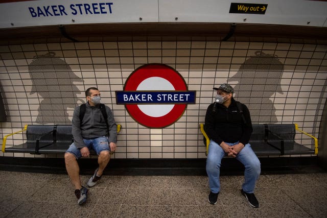 Two men in face masks sit in front of Sherlock Holmes artwork in Baker Street Tube station (Victoria Jones/PA)