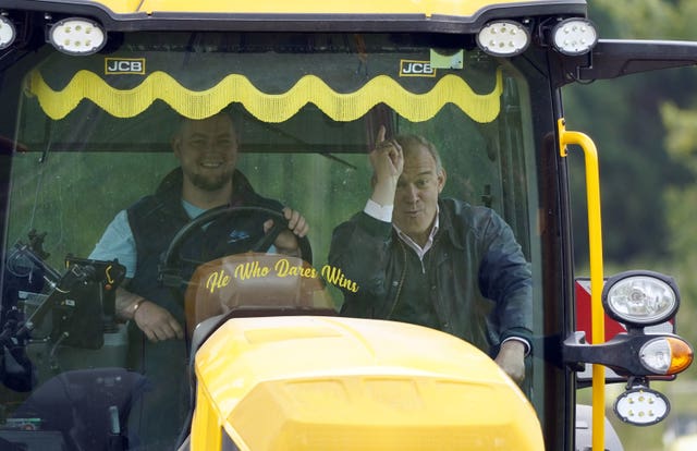 Liberal Democrat leader Sir Ed Davey points upwards as he is driven in a JCB Fastrac, which has 'He Who Dares Wins' written on the bottom of the windscreen, during a visit to Owl Lodge in Lacock, Wiltshire