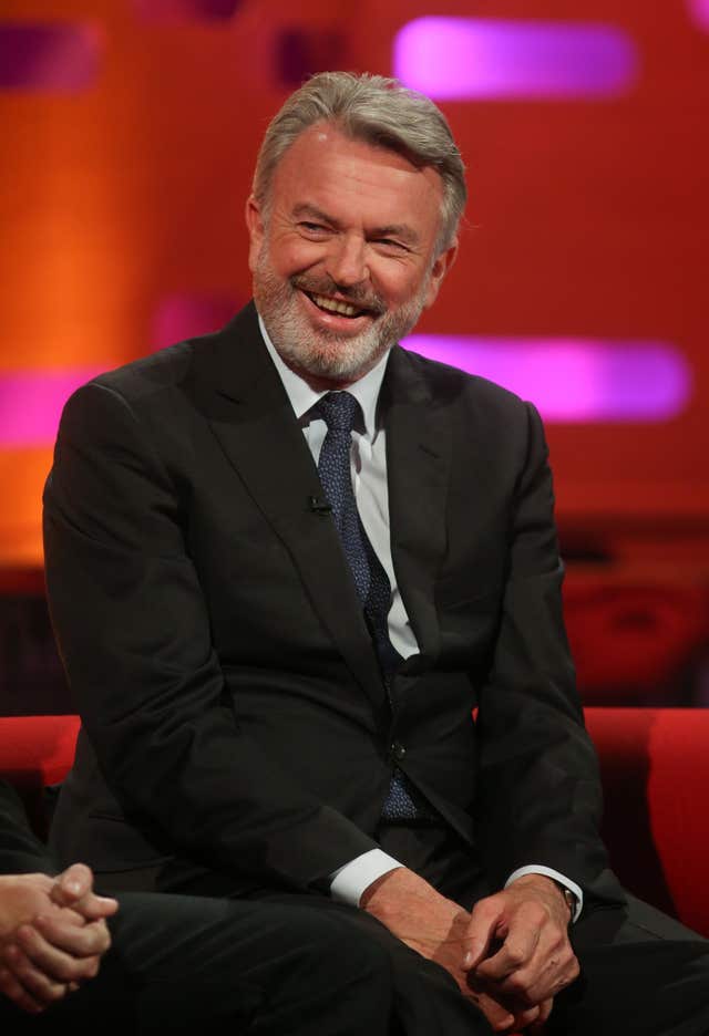 Sam Neill smiling while on a red sofa