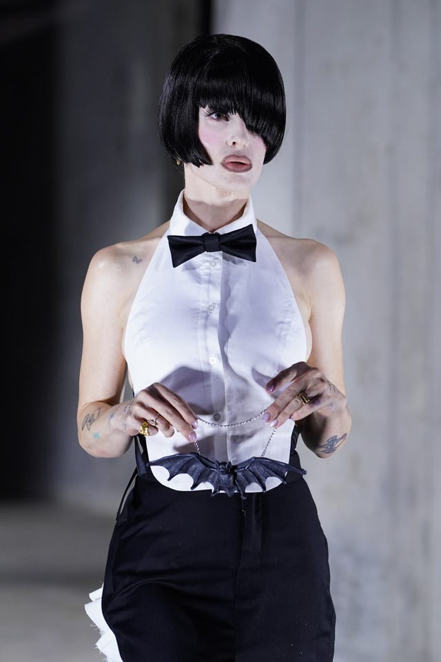 Kelly Osbourne holding a bat necklace wearing a white waistcoat, bow tie and black skirt