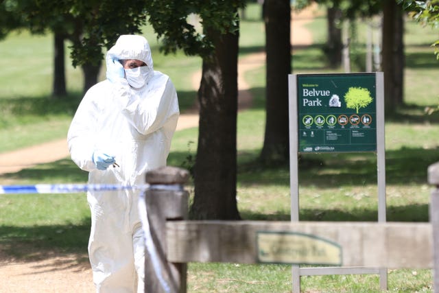 An officer in a white forensic suit standing by a line of police tape in a park
