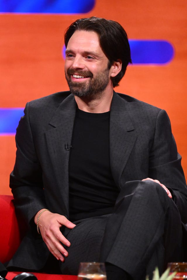 Sebastian Stan smiling while sitting on a sofa, dressed in suit and T-shirt