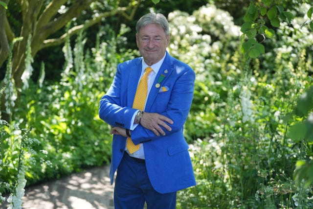 Alan Titchmarsh standing in a garden with his arms folded wearing a blue suit and a yellow tie