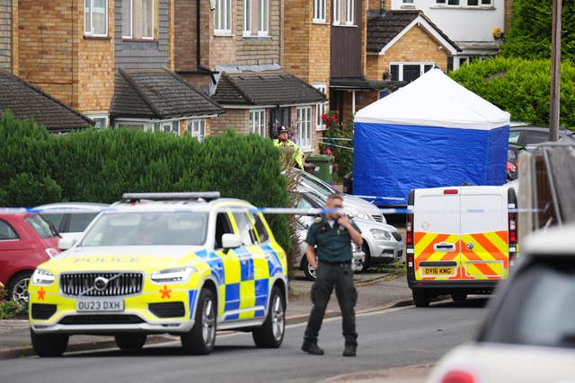 A police officer stands beside a car and blue forensic tent behind police cordon tape