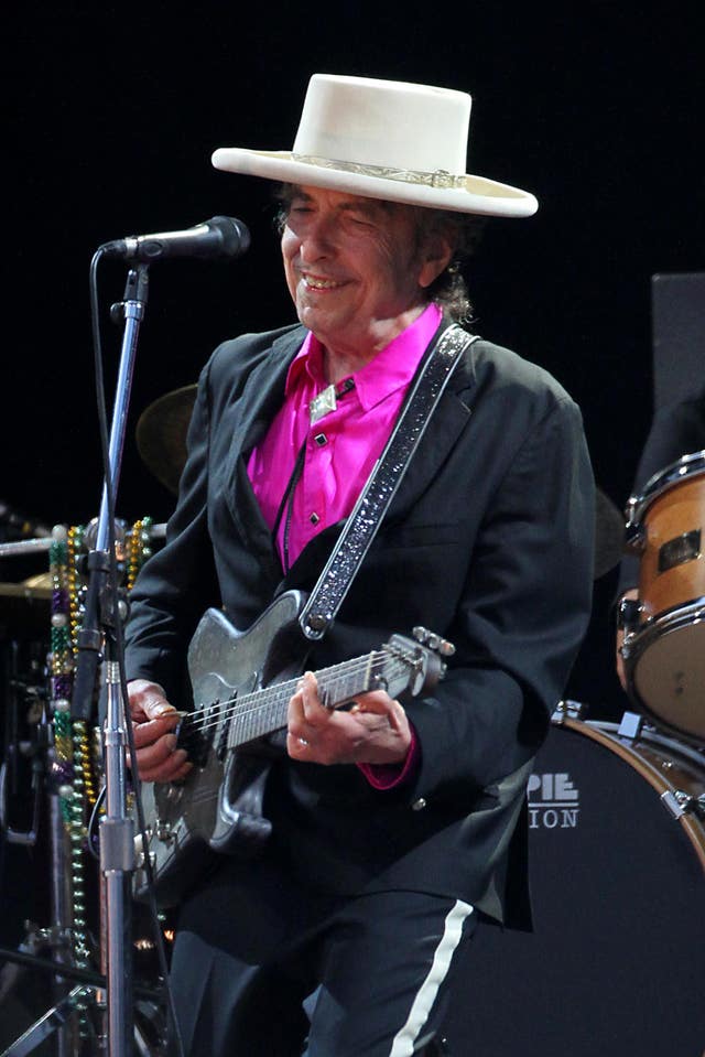 Bob Dylan, wearing a white hat, pink shirt and dark suit,  smiles while playing the guitar during a performance at the Hop Farm Festival