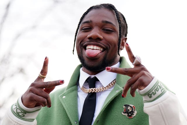 Noah Lyles, in a green and white varsity jacket, sticks out his tongue as he gestures to the camera