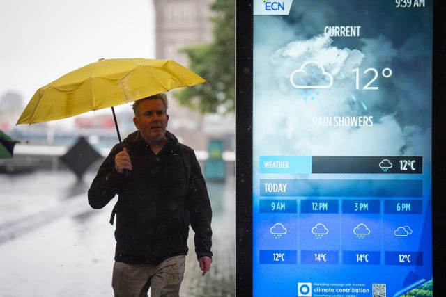 A man walking with a yellow umbrella over his head while a digital information board in the foreground displays a rainy weather forecast