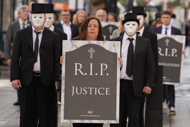 People in white masks and dark suits with a woman holding a gravestone-shaped sign reading RIP Justice