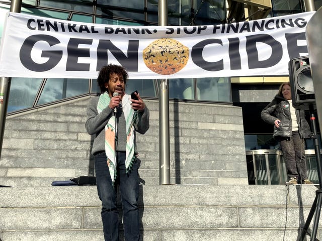 Darragh Adelaide speaking into a microphone, below a banner reading 'Central Bank: Stop financing genocide'