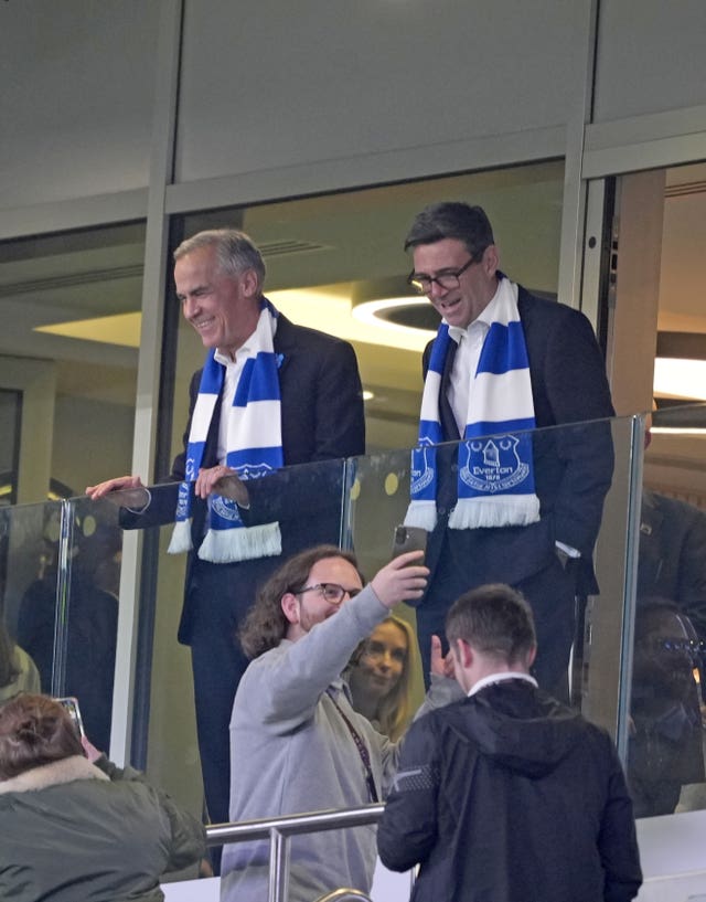 Mark Carney and Andy Burnham in a box at the Hill Dickinson Stadium while a fan in the foreground takes a selfie
