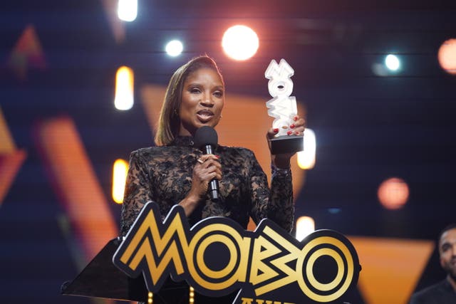 Denise Lewis speaking into a microphone she is holding in one hand as she grasps a Mobo Award with her other hand