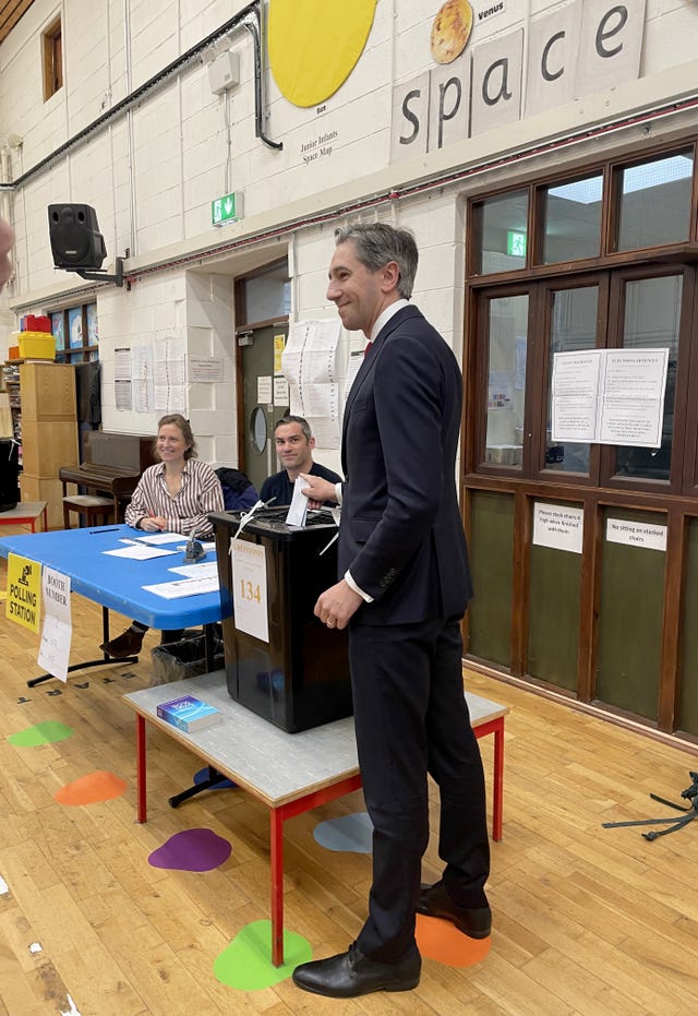 Taoiseach Simon Harris in a school gym posing for photographs after casting his vote. He stands next to the black box where votes are deposited.