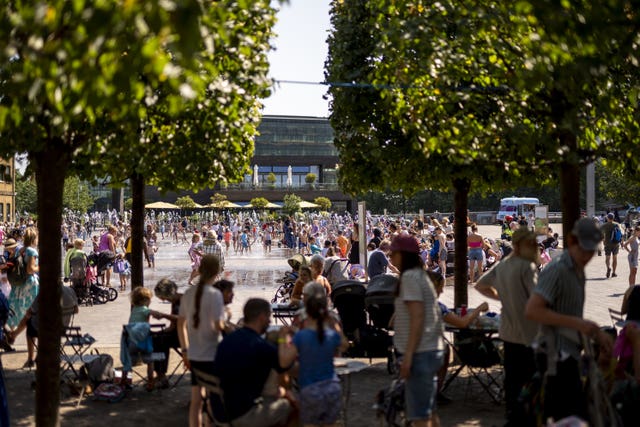 Crowds of people shaded by trees look out at people having fun around a fountain