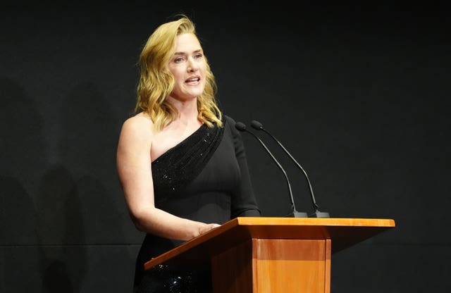Kate Winslet in a black dress speaking from behind a lectern
