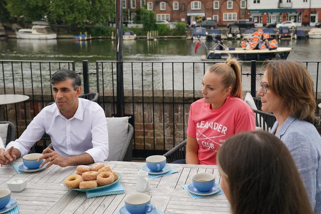 Rishi Sunak sitting down at a table speaking to rowing club members while Liberal Democrat supporters pass in the river behind him on a boat