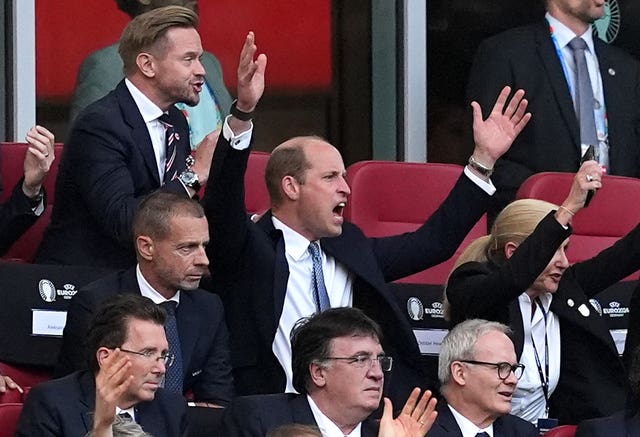 A group of people in the stands, the Prince of Wales is in the centre with his hands in the air, captured mid-shout