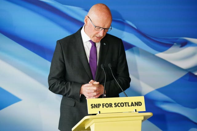 John Swinney clasps his hands while standing behind a podium that says SNP for Scotland