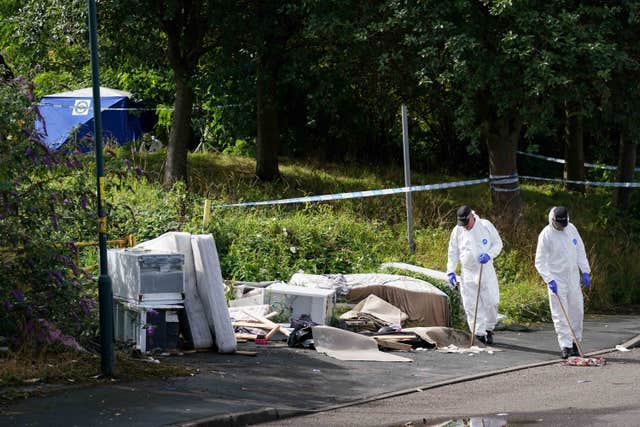 Two forensic officers search through rubbish left on the side of a woodland area, a forensic tent in the background