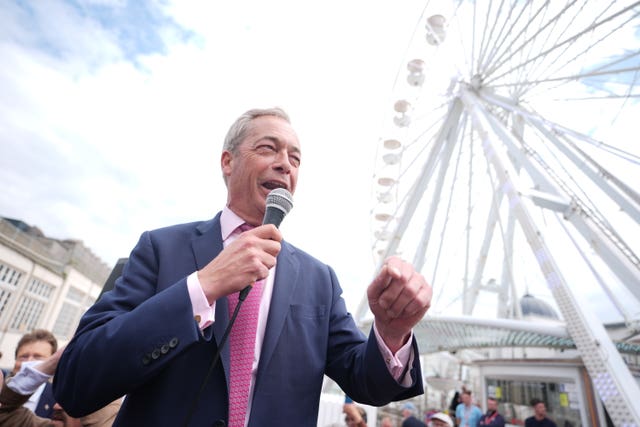 Nigel Farage standing in front of a fairground big wheel and speaking into a microphone