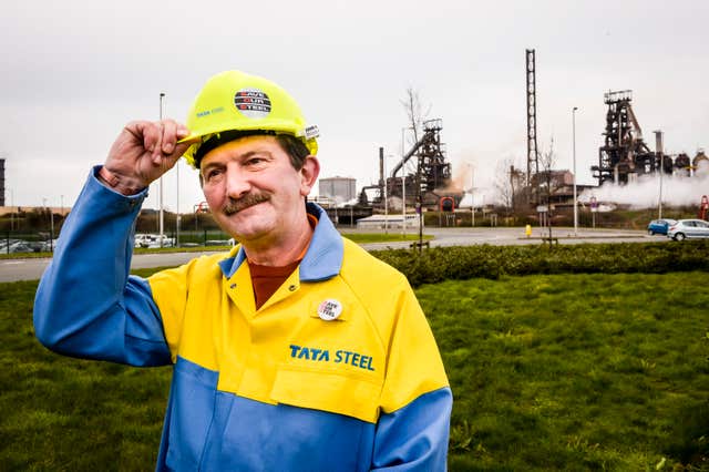 A Tata Steel worker dressed in a red and blue boiler suit touches his hard hat, standing in front of the steelworks