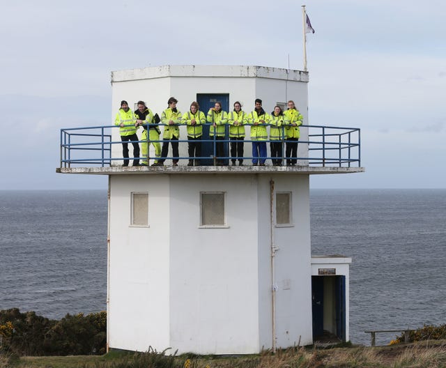 A group of young people wearing hi-vis jackets standing at the top of a watchtower on a clifftop