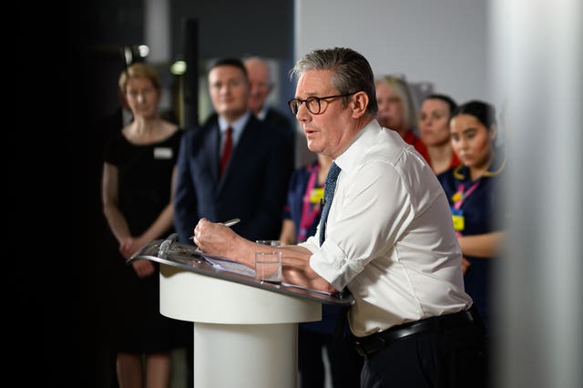 Sir Keir Starmer wearing a white shirt with his sleeves rolled up leaning on a lectern while the Health Secretary and NHS staff look on