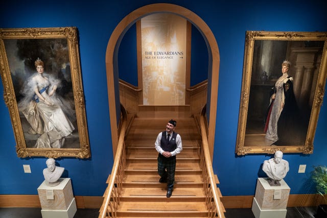 A man walking down a staircase, flanked by large paintings of Queen Alexandra and Queen Mary
