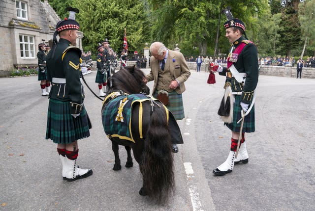 The King, in a beige jacket and tartan kilt, pats the head of a brown pony 