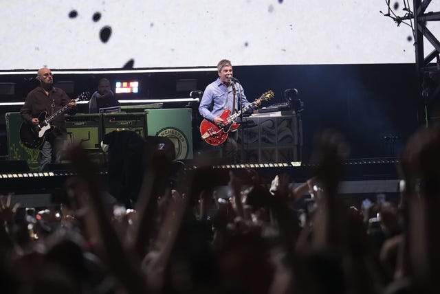 Noel Gallagher performing on stage in the background with fans waving their hands in the air in the foreground