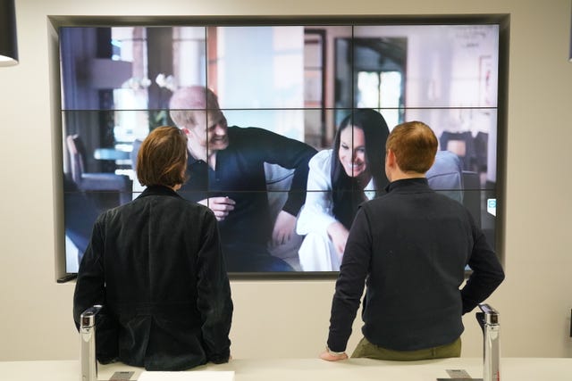 The backs of two people watching a large screen showing the Duke and Duchess of Sussex smiling