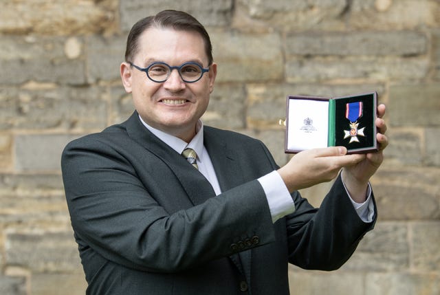 Professor Paul Mealor smiles while holding up his RVO medal