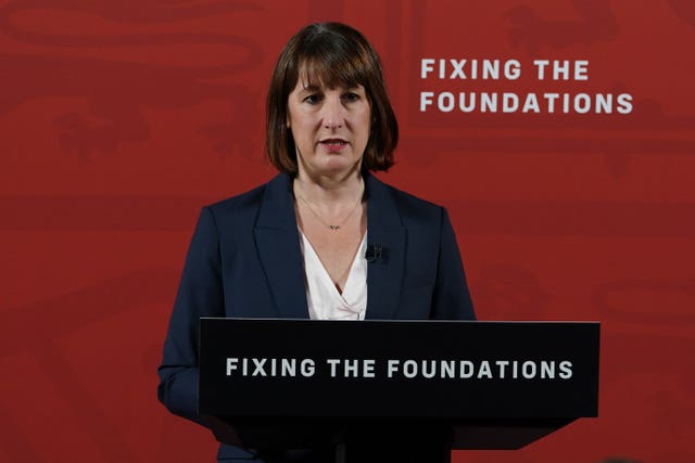 Rachel Reeves stands at a lectern with the slogan "Fixing the Foundations" written on it. The same slogan is written on a red wall behind her