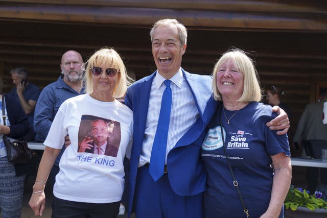 Nigel Farage standing in the middle of two female supporters wearing Reform UK T-shirts, including one featuring a picture of Farage smoking a cigarette with the words 'The King' beneath it