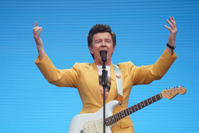 Rick Astley gestures with hands as he performs on stage with a guitar