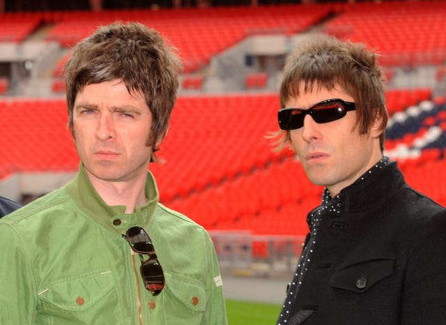 Noel and Liam Gallagher posing together at Wembley Stadium, with Noel in a green jacket and Liam in a black jacket and wearing dark sunglasses