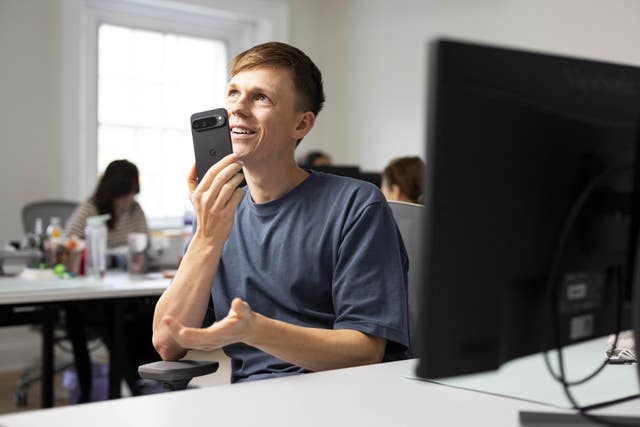 YouTuber Caspar Lee holding a phone to his mouth while sat on a desk behind a computer screen