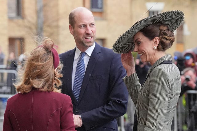 Kate holding on to her hat as she and William chat to a woman, who has her back to the camera, outside